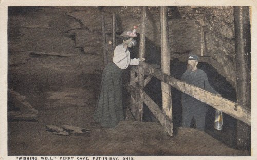 A woman in a long dress leans over a wooden railing at a cave wishing well, while a man in a uniform holds a lantern.