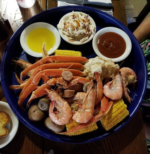 A large seafood platter with shrimp, crab legs, corn, potatoes, coleslaw, and dipping sauces on a blue plate.