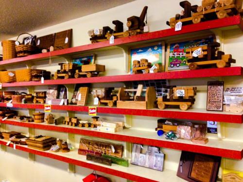 A display of various wooden toys and crafts on red shelves, featuring trucks, baskets, and decorative items.