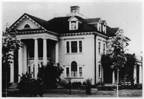 A large, historic house with columns, a steep roof, and surrounding trees, captured in black and white.