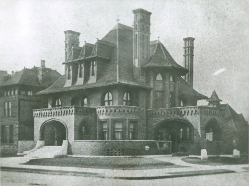 Historic mansion with ornate architecture, featuring towers, arches, and a distinctive roof, set on a street corner.