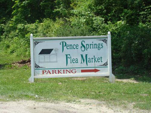 Sign for Pence Springs Flea Market with a parking arrow pointing to the left, surrounded by greenery.