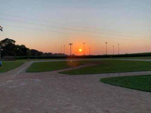 A serene baseball field at sunset, with lights on and a warm glow in the sky.