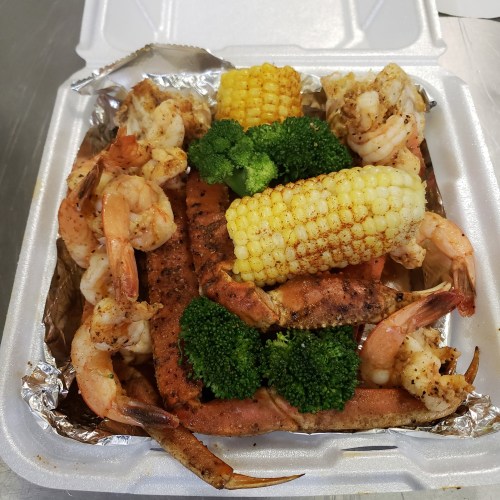 A takeout container filled with shrimp, crab, corn on the cob, and broccoli, all seasoned and cooked.