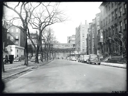 Black and white photo of a street with vintage cars, trees, and an elevated train track in the background.
