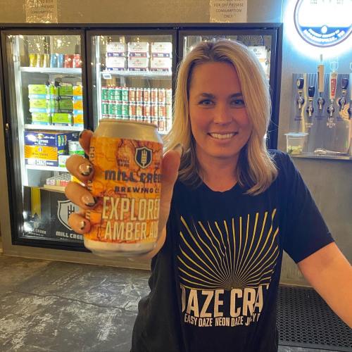 A woman holds a can of Mill Creek Brewing's Explore Amber Ale, smiling in front of a cooler filled with drinks.