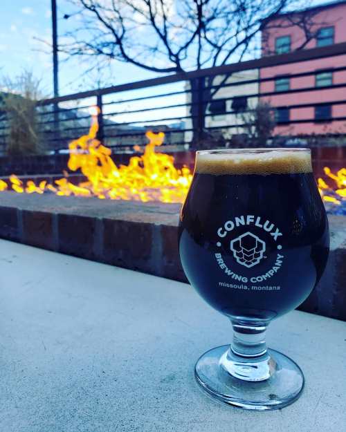 A glass of dark beer from Conflux Brewing Company sits in front of a fire pit, with trees and buildings in the background.