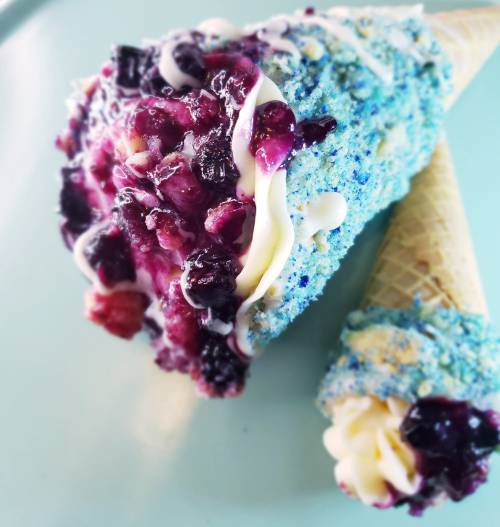 Two colorful ice cream cones topped with vibrant berries and creamy swirls, resting on a light blue plate.