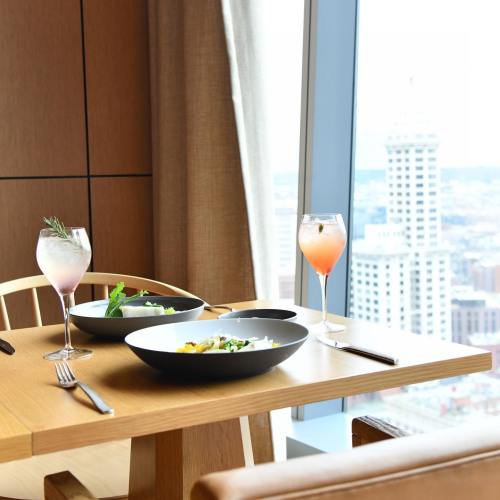 A dining table with two cocktails and bowls of salad, overlooking a city skyline through a large window.