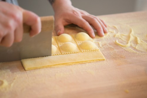 A chef's hands cutting fresh pasta into ravioli shapes on a wooden surface dusted with flour.
