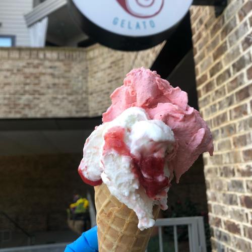 A close-up of a cone filled with pink and white gelato, with a blurred shop sign in the background.