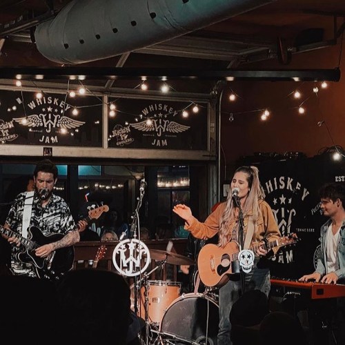 A live music performance at a venue with string lights, featuring a singer with a guitar and a band on stage.