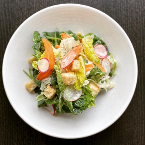 A fresh salad with greens, tomatoes, radishes, croutons, and dressing in a white bowl on a dark surface.