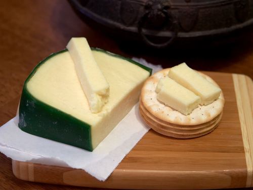 A wedge of cheese with a green rind next to a stack of crackers topped with cheese slices on a wooden board.