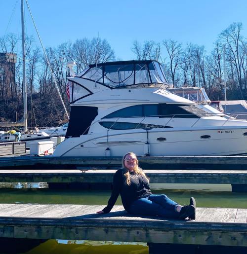A woman sits on a dock by a marina, smiling with a yacht in the background under a clear blue sky.