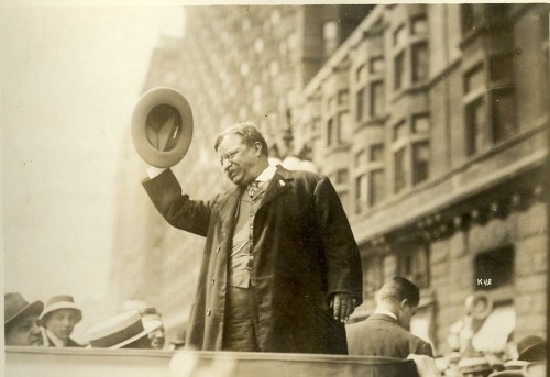 A historical figure in a suit and hat waves to a crowd from a platform, surrounded by onlookers in early 20th-century attire.