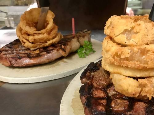 Two grilled steaks on plates, one topped with onion rings and the other with a crispy onion tower, garnished with parsley.