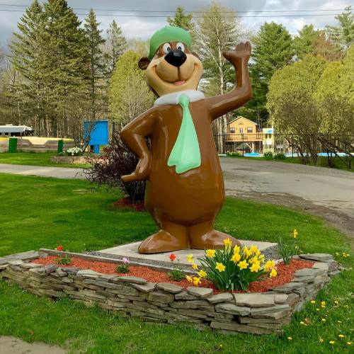 A large, cheerful bear statue wearing a green hat and scarf waves, surrounded by flowers and trees.