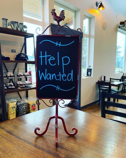 A decorative chalkboard sign reading "Help Wanted" stands on a table in a cozy café setting.