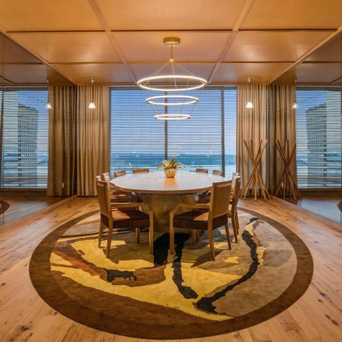 A modern dining room with a round table, wooden chairs, a circular rug, and large windows overlooking a scenic view.