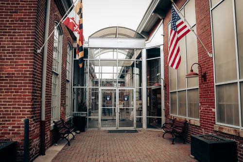 Entrance of a brick building with large glass doors, featuring American and Maryland flags on either side.