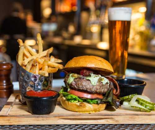 A juicy burger with lettuce, tomato, and sauce, served with fries, ketchup, pickles, and a pint of beer on a wooden board.