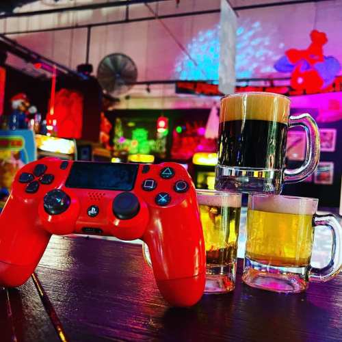 A red gaming controller sits on a table next to two small beer mugs and a large glass of dark beer in a colorful setting.