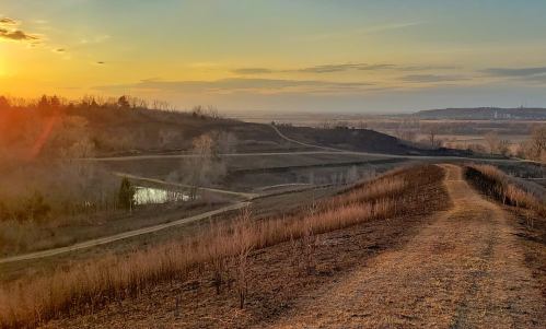 A scenic landscape at sunset, featuring rolling hills, a winding path, and a calm pond in the foreground.