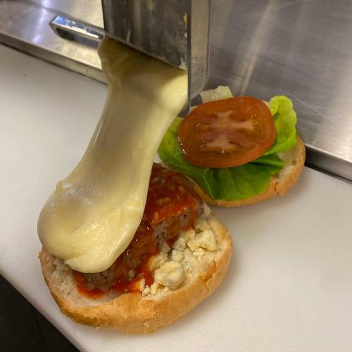 Melted cheese being poured over a burger patty on a bun, topped with lettuce and tomato.