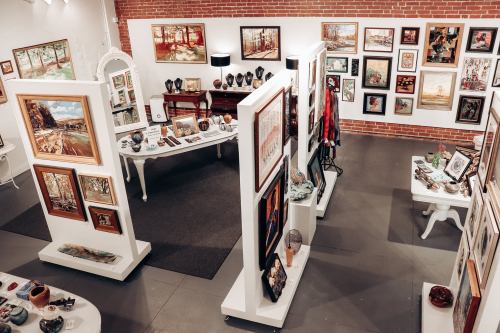 An art gallery featuring various paintings and sculptures displayed on walls and tables in a bright, open space.