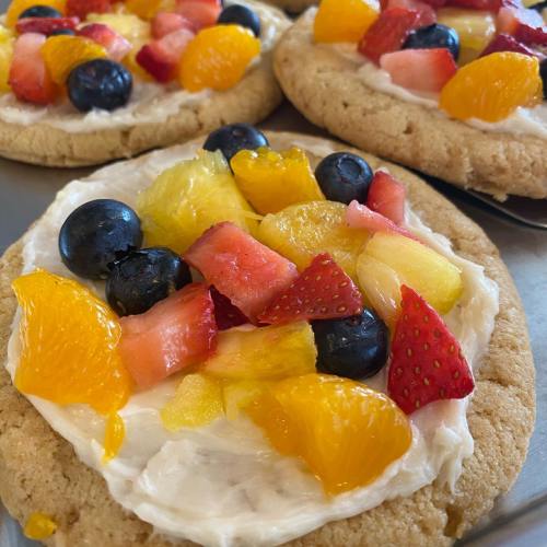 Close-up of large cookies topped with cream cheese frosting and a colorful assortment of fresh fruits.