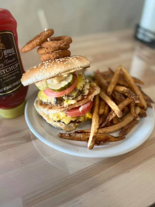 A towering cheeseburger with pickles, tomatoes, and onion rings, served with a side of fries and ketchup.