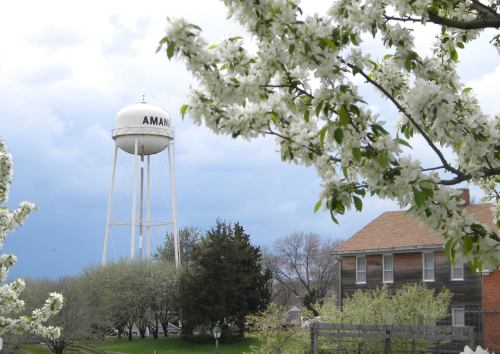 A white water tower labeled "Amana" stands among blooming trees and a historic building under a cloudy sky.