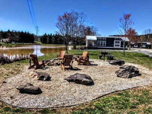 A cozy outdoor seating area with Adirondack chairs around a fire pit, near a pond and surrounded by trees.
