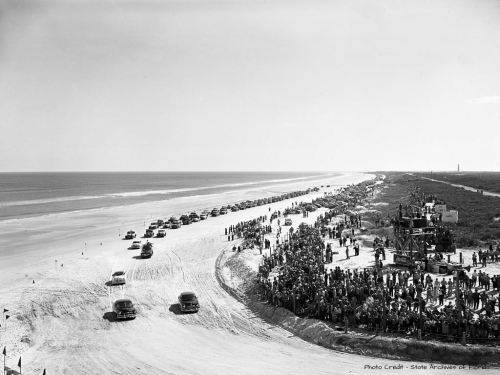 A historic black-and-white photo of cars driving on a beach, with a large crowd watching from the shore.