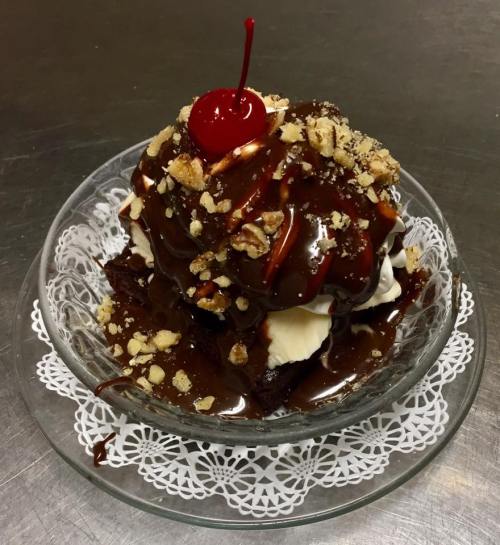 A decadent dessert with chocolate sauce, whipped cream, nuts, and a cherry on top, served in a glass bowl.