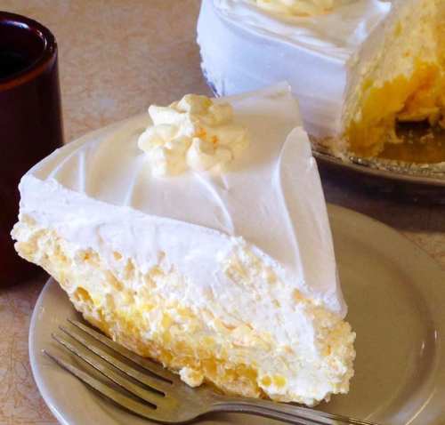 A slice of creamy lemon pie topped with whipped cream, served on a plate with a fork and a cup of coffee in the background.