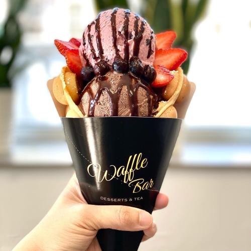 A hand holding a waffle cone filled with ice cream, strawberries, and chocolate drizzle, labeled "Waffle Bar."
