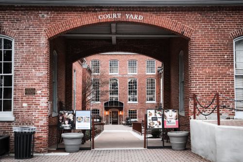 A brick archway leads to a courtyard with signs and a view of a building in the background.