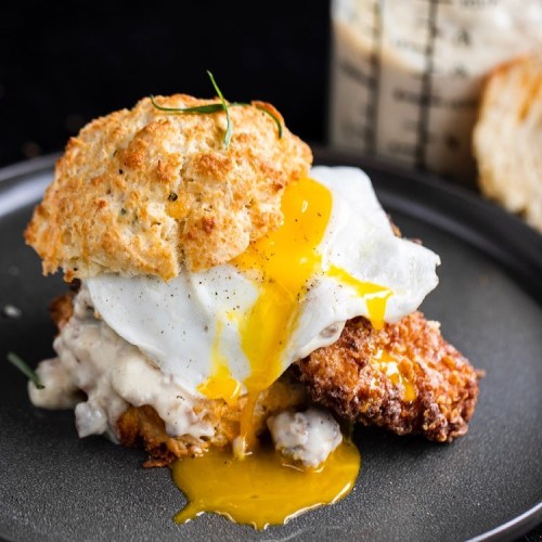 A biscuit sandwich with fried chicken, topped with a runny egg and creamy gravy, served on a dark plate.