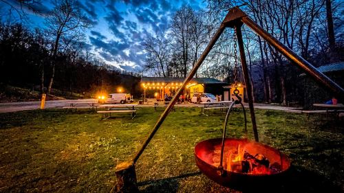 A campfire setup in the foreground with a cozy cabin and string lights in the background, surrounded by trees at dusk.