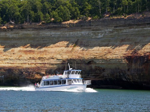 A white tour boat glides through blue water near a colorful, layered cliffside under a clear sky.