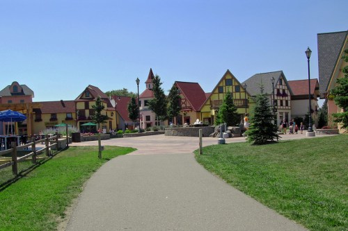 A scenic pathway leads through a charming village with colorful buildings and greenery under a clear blue sky.