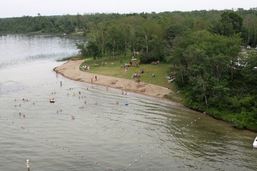 A sandy beach by a lake with people swimming and relaxing on the shore, surrounded by trees and green grass.
