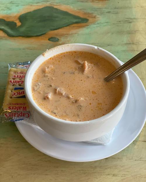 A bowl of creamy soup with chunks of meat, served with a small packet of crackers on a white plate.