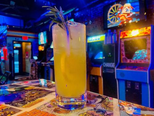 A refreshing cocktail with a sprig of rosemary sits on a colorful tabletop, with arcade machines in the background.