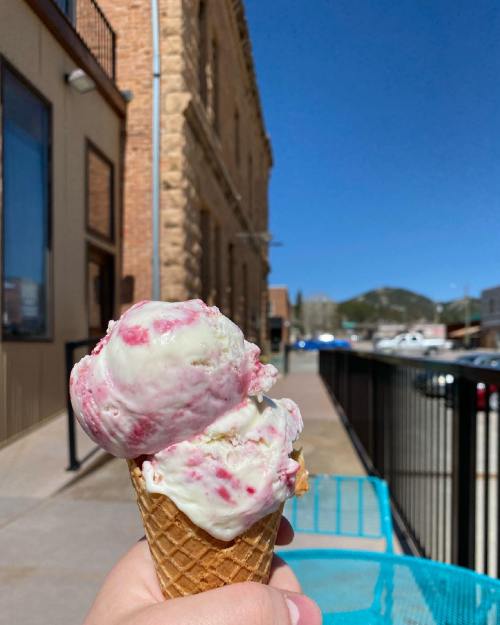 A hand holds a cone with two scoops of pink and white ice cream, with a sunny street and buildings in the background.