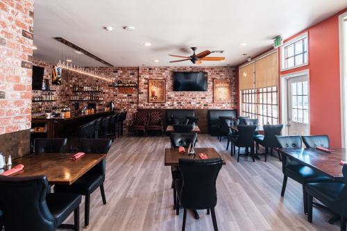 Interior of a restaurant featuring brick walls, wooden tables, black chairs, and a bar area with a TV.