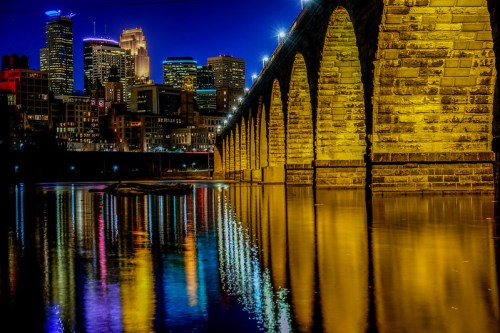 A stone bridge arching over a river, reflecting city lights and skyscrapers at night.