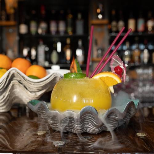 A vibrant cocktail in a shell-shaped glass, garnished with fruit and smoke, set against a bar backdrop.